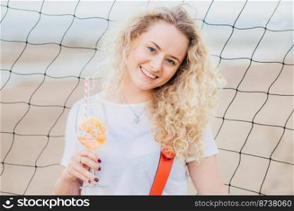 Horizontal shot of cheerful glad blonde female dressed in casual clothes, holds fresh orange cocktail, smiles positively, poses against tennis net background at beach. Woman with summer beverage