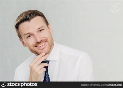 Horizontal shot of cheerful male with stubble, holds chin, looks positively at camera, dressed in formal shirt with tie, listens something pleasant, isolated over white background. Man employee