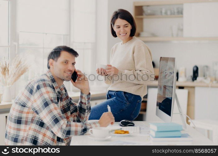 Horizontal shot of cheerful man has telephone conversation, discusses financial contract agreement, dressed in checkered shirt, writes down information, happy woman holds mug of drink, leans at table