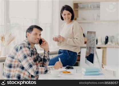 Horizontal shot of cheerful man has telephone conversation, discusses financial contract agreement, dressed in checkered shirt, writes down information, happy woman holds mug of drink, leans at table
