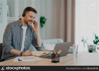 Horizontal shot of cheerful man participates in self improvement webinar wears checkered shirt communicates online by video call uses headset and laptop computer studies online poses at desktop