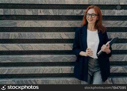 Horizontal shot of cheerful positive young redhead woman dressed formally drinks takeaway coffee holds digital tablet and notepad dressed in formal clothes stands near marbe wall copy space away. Woman dressed formally drinks takeaway coffee holds digital tablet and notepad