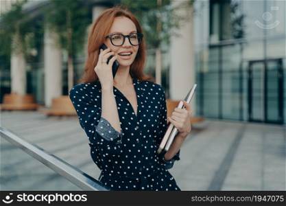 Horizontal shot of cheerful redhead European woman talks on modern mobile phone has glad expression smiles broadly holds laptop and diary walks outside against blurred background during daytime. Cheerful redhead European woman talks on modern mobile phone has glad expression