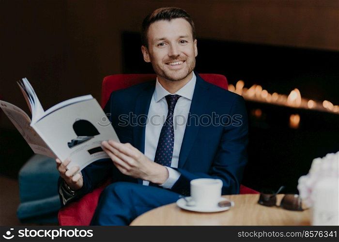 Horizontal shot of cheerful successful young businessman being in good mood, smiles positively, holds journal in hands, drinks aromatic coffee during work break, dressed in formal luxury suit