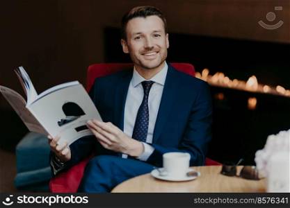 Horizontal shot of cheerful successful young businessman being in good mood, smiles positively, holds journal in hands, drinks aromatic coffee during work break, dressed in formal luxury suit