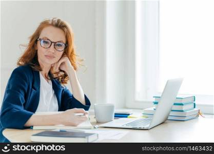 Horizontal shot of confident successful redhead female business owner sits at cabinet, works on laptop computer, writes main theses in notebook, has coffee break, poses in spacious office room