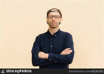 Horizontal shot of confident young handsome male worker, wears cap and shirt, keeps arms folded, has delighted expression, listens inforamtion attentively, poses against beige studio background