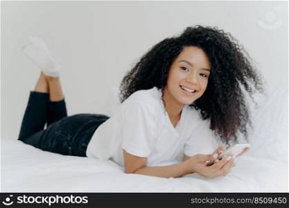 Horizontal shot of curly ethnic woman with pleasant smile, lies on comfortable bed, raises legs, uses modern cellphone, reads text message, enjoys high speed internet connection. People and bed time