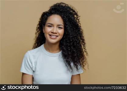 Horizontal shot of curly haired woman with healthy skin, toothy smile, enjoys being photographing, wears casual white t shirt, isolated on beige background. People, emotions and lifestyle concept