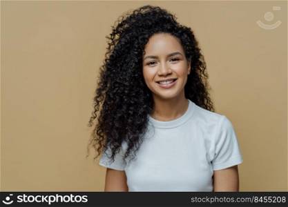 Horizontal shot of curly haired woman with healthy skin, toothy smile, enjoys being photographing, wears casual white t shirt, isolated on beige background. People, emotions and lifestyle concept
