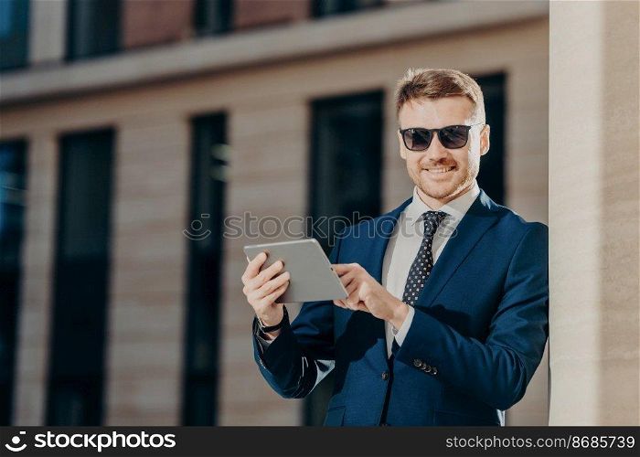 Horizontal shot of delighted young businessman monitors business rate of companies via touch pad in internet, wears sunglasses and formal clothes, has happy expression and pleasant appearance