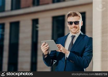 Horizontal shot of delighted young businessman monitors business rate of companies via touch pad in internet, wears sunglasses and formal clothes, has happy expression and pleasant appearance