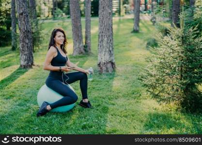 Horizontal shot of European brunette woman dressed in cropped top and leggings, sits on fitness ball, uses smartphone and listens music in earphones, holds bottle of fresh water, poses in forest
