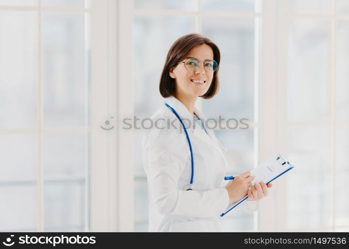 Horizontal shot of female doctor fills up medical form at clipboard, stands indoor, wears round glasses, white gown and stethoscope. General practitioner writes down notes, consults patients