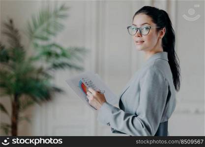 Horizontal shot of female strartuper analyzes business information, holds paper documents with diagrams and graphics, looks confidently at camera, wears transparent glasses and formal clothes