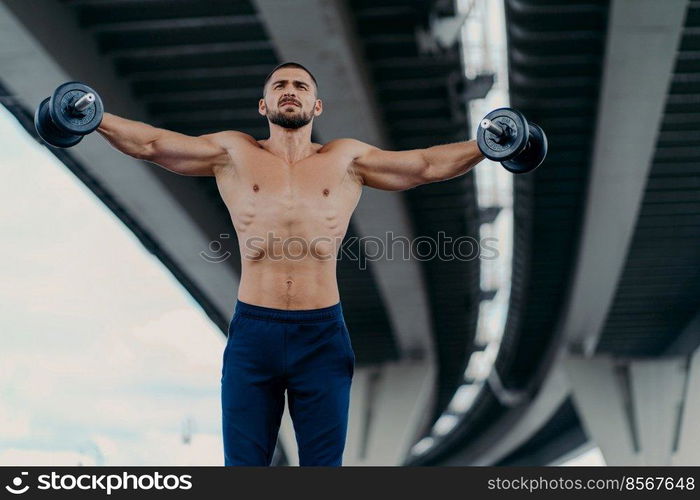 Horizontal shot of fit muscular bearded man works out with heavy weights trains muscles and raises arms being professional bodybuilder demonstrates determination stands powerful under bridge