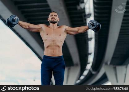 Horizontal shot of fit muscular bearded man works out with heavy weights trains muscles and raises arms being professional bodybuilder demonstrates determination stands powerful under bridge