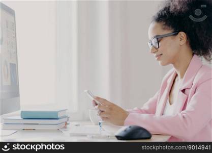 Horizontal shot of focused curly haired female office worker holds mobile phone, searches information in internet, wears optical glasses and elegant suit, computer monitor in front, does paper work