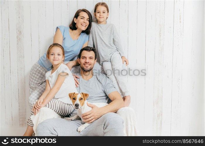Horizontal shot of friendly family pose together against white background  two little sisters, father, mother and their pet. Happy parents and their female children. Family of four. Parenthood