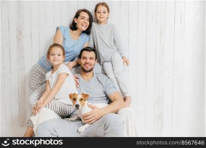 Horizontal shot of friendly family pose together against white background  two little sisters, father, mother and their pet. Happy parents and their female children. Family of four. Parenthood