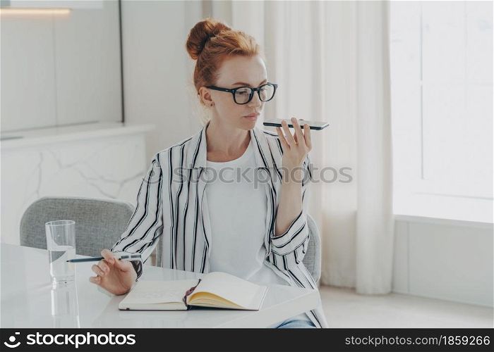 Horizontal shot of ginger woman sits at table makes notes in notepad uses virtual assistant to record message or make call sits at desktop dressed casually poses against home interior. Technology. Woman sits at table makes notes in notepad uses virtual assistant to record message or make call