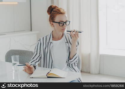 Horizontal shot of ginger woman sits at table makes notes in notepad uses virtual assistant to record message or make call sits at desktop dressed casually poses against home interior. Technology. Woman sits at table makes notes in notepad uses virtual assistant to record message or make call