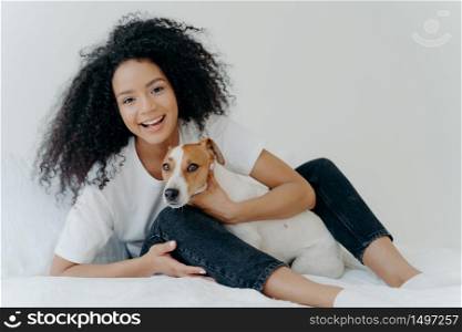 Horizontal shot of glad Afro woman rests in bed with dog, have playful mood, pose together in bedroom against white background. Girl relaxes at home with jack russell terrier. Sweet funny moment