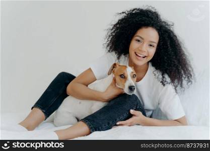 Horizontal shot of glad Afro woman rests in bed with dog, have playful mood, pose together in bedroom against white background. Girl relaxes at home with jack russell terrier. Sweet funny moment