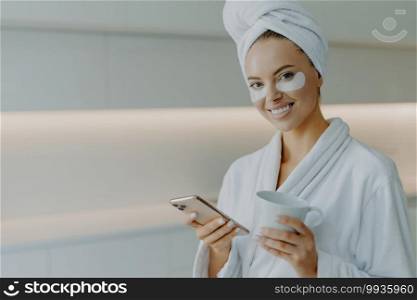 Horizontal shot of glad beautiful womanwith healthy skin cares about undereye skin applies moisturising patches poses in bathrobe uses mobile phone for surfing social networks drinks coffee.