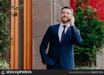 Horizontal shot of glad male entrepreneur speaks via smart phone, looks into distance, wears formal black suit, white shirt and tie, stands outdoor, waits for business partner near cafe or restaurant