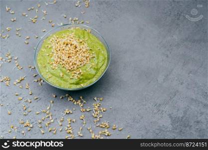 Horizontal shot of glass bowl with fresh vegetetable green smoothie containig much vitamins, buckwheat sprouts around and on top. Dark background. Copy space