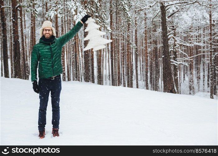 Horizontal shot of good looking bearded male looks with happy expression, demonstrates artificial fir tree which he bought, walks on snowy weather in beautiful forest. Happiness concept