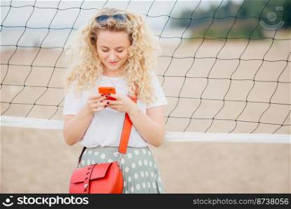 Horizontal shot of good looking female with curly hair, holds modern cell phone, texts messages to friend, stands near tennis net, strolls on beach. Beautiful woman surfes internet on cellular