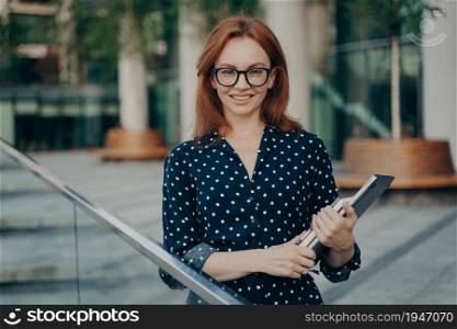 Horizontal shot of good looking ginger prosperous businesswoman poses outdoor in stylish outfit carries necessary items for work looks directly at camera. People occupation and lifestyle concept. Prosperous businesswoman poses outdoor in stylish outfit carries necessary items for work