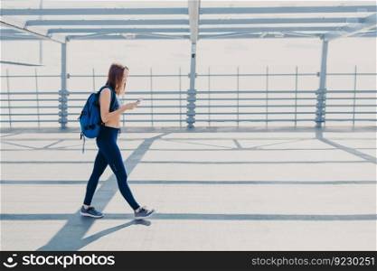 Horizontal shot of good looking sportswoman dressed in casual outfit, carries rucksack, being addicted to modern technologies, always in touch, listens favourite song. Active lifestyle concept