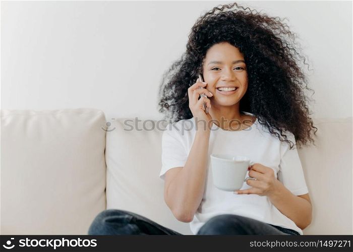 Horizontal shot of good looking woman has distant conversation via mobile phone, solves informal issues while talking, sits on sofa in living room, drinks tea, has broad smile, shows white teeth