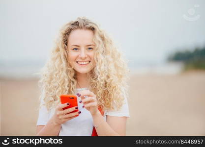 Horizontal shot of good looking young blonde female has curly hair, holds orange smart phone, messages with friends, poses against blurred beach background, rests in tropical country with lover