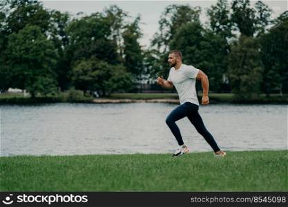 Horizontal shot of handsome active bearded man goes running being photographed in motion, runs against river and beautiful nature, gets ready for marathon. Sport, wellness and workout concept