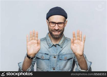 Horizontal shot of handsome bearded male shows palms at camera, demonstrates refusal sign, looks with mysterious expression into camera, has thick beard and mustache, isolated on white background