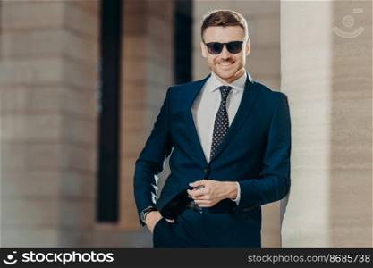Horizontal shot of handsome cheerful bearded young male manager with confident cheerful expression, keeps hand in pocket, poses against office interior. People, business and elegance concept