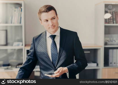 Horizontal shot of handsome male manager in stylish clothes, uses smart phone for checking information about entrepreneurship, reads news in web page, connected to wireless internet, stands at office
