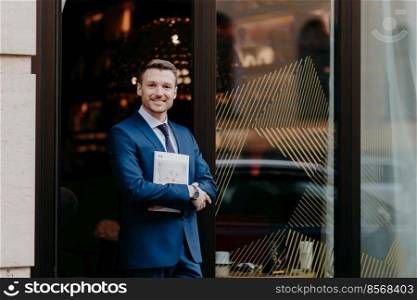 Horizontal shot of handsome male with cheerful expression, dressed in formal suit, holds magazine, stands against cafe background, going to have lunch during work break. Successful businessman