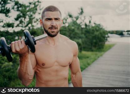 Horizontal shot of handsome muscular man lifts barbell outdoor, has athletic torso, gets ready for weight lifting training, has muscular arms, poses against street background. Developing strength