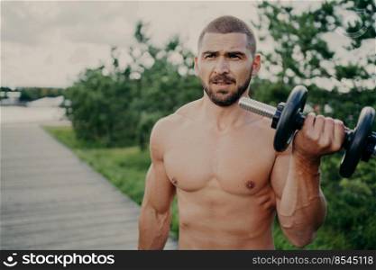 Horizontal shot of handsome muscular man lifts barbell outdoor, has athletic torso, gets ready for weight lifting training, has muscular arms, poses against street background. Developing strength