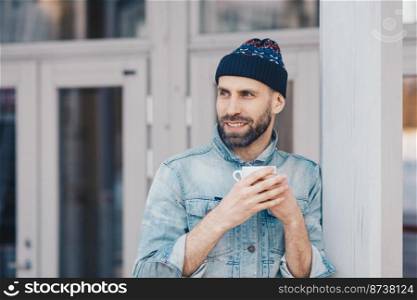 Horizontal shot of handsome smiling male looks happily away, wears hat and jean jacket, holds mug of coffee, notices something pleasant, rests at home. People, lifestyle and recreation concept