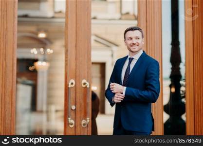 Horizontal shot of handsome successful male executive worker stands near doors of office building, goes on business meeting, waits for colleague, looks positively aside. People and business concept