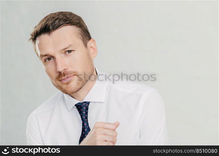 Horizontal shot of handsome young male entrepreneur in formal white shirt, looks directly at camera, listens attentively his interlocutor, talk on business theme, isolated over white background