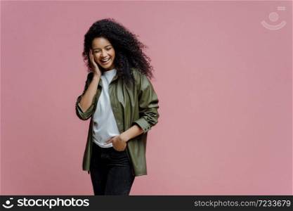 Horizontal shot of happy African American woman keeps hand in pocket, laughs as hears funny anecdote, spends free time in merry company, concentrated down, isolated on pink background, copy space