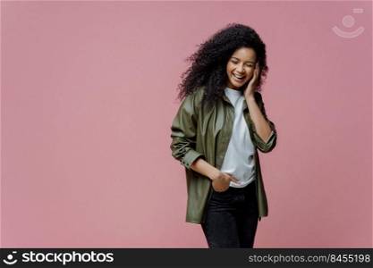 Horizontal shot of happy African American woman keeps hand in pocket, laughs as hears funny anecdote, spends free time in merry company, concentrated down, isolated on pink background, copy space