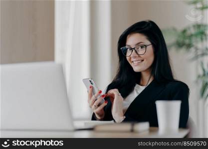 Horizontal shot of happy brunette European woman messages on modern smartphone, gives online consultation, smiles positively, wears spectacles and formal suit, sits in front of opened laptop.